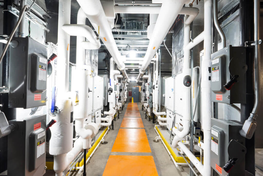 Interior view of a geothermal water source heat pump system at Louisville Muhammad Ali International Airport, featuring rows of insulated pipes, electrical boxes, and HVAC units along a brightly lit corridor.