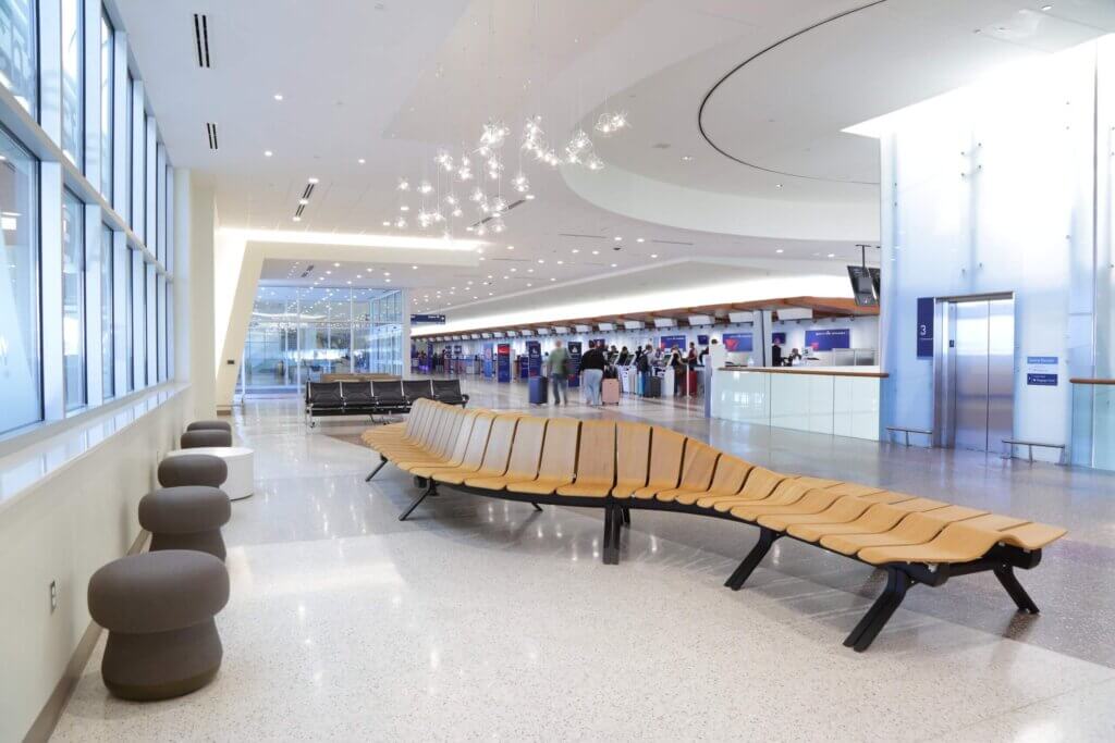 Modern airport terminal with sleek wooden seating, contemporary lighting, and check-in counters in the background. The area is clean, spacious, and well-lit with natural light streaming through large windows.